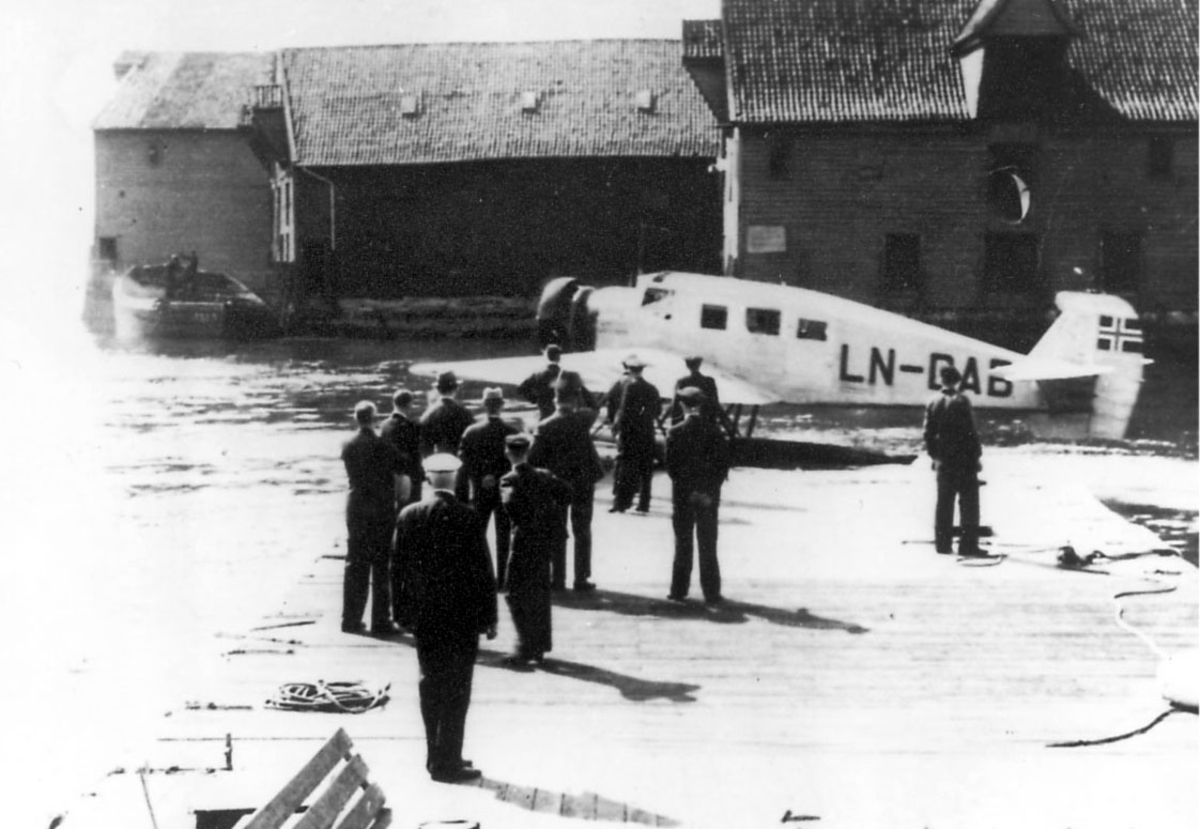 Ett fly på bakken, Junkers Ju W 34 LN-DAB  "Ternen".Flere personer ved flyet. Brygger - bygninger i bakgrunnen