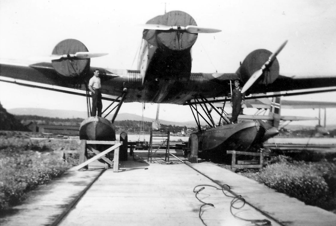 Sjøflyhavn, ett fly på bakken, Junkers Ju 52/3mg3e. Presenninger over motorene. to personer ved flyet. Litt av ett annet fly sees i bakgrunnen.