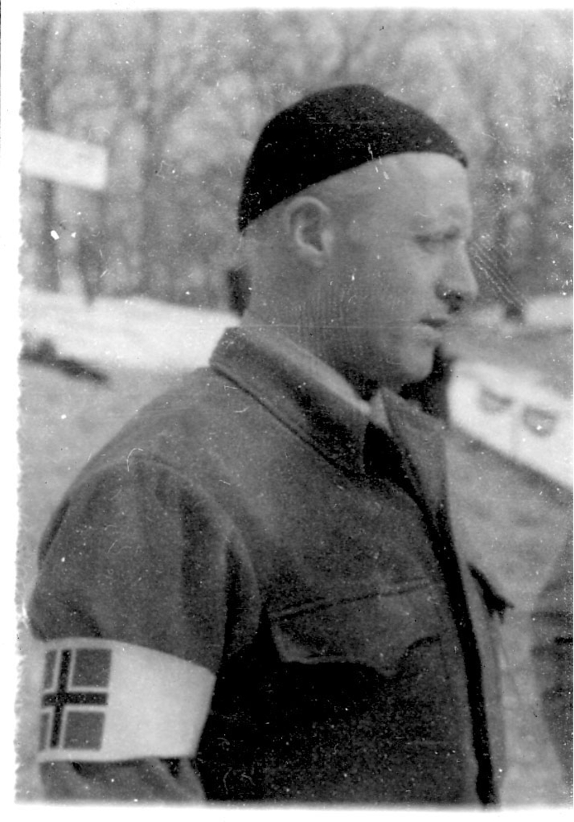 Portrett, en person i militæruniform. Hvitt bånd med det norske flagg på armen. Tatt utendørs.