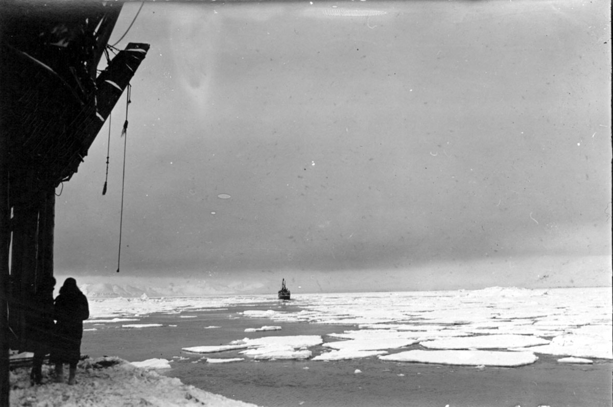 Fra kai (heiseutstyr?). 2 personer. Fartøy/båt utpå havet, i bakgrunnen. Hav, små isflak. Snø.