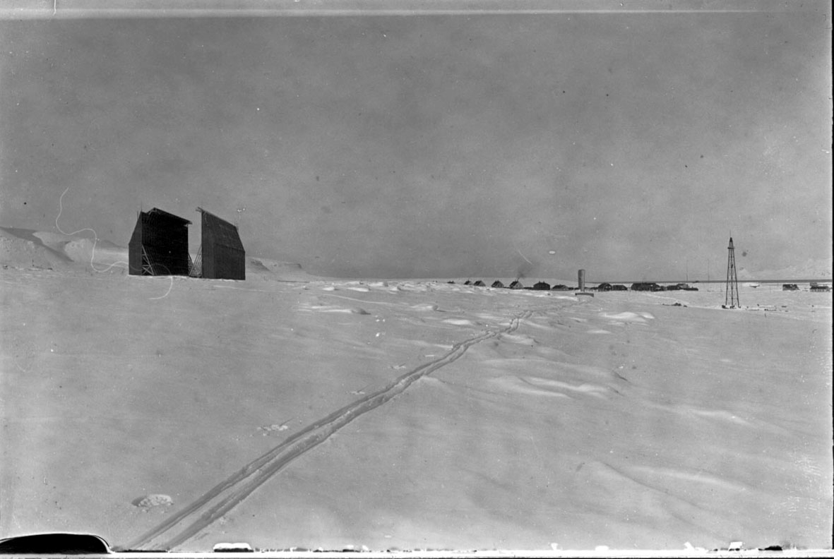 Luftskipshangaren t.v., fortøyningsmast t.h. Flere bygninger, husklynge, i bakgrunnen. Skispor i forgrunnen, snø på bakken. 