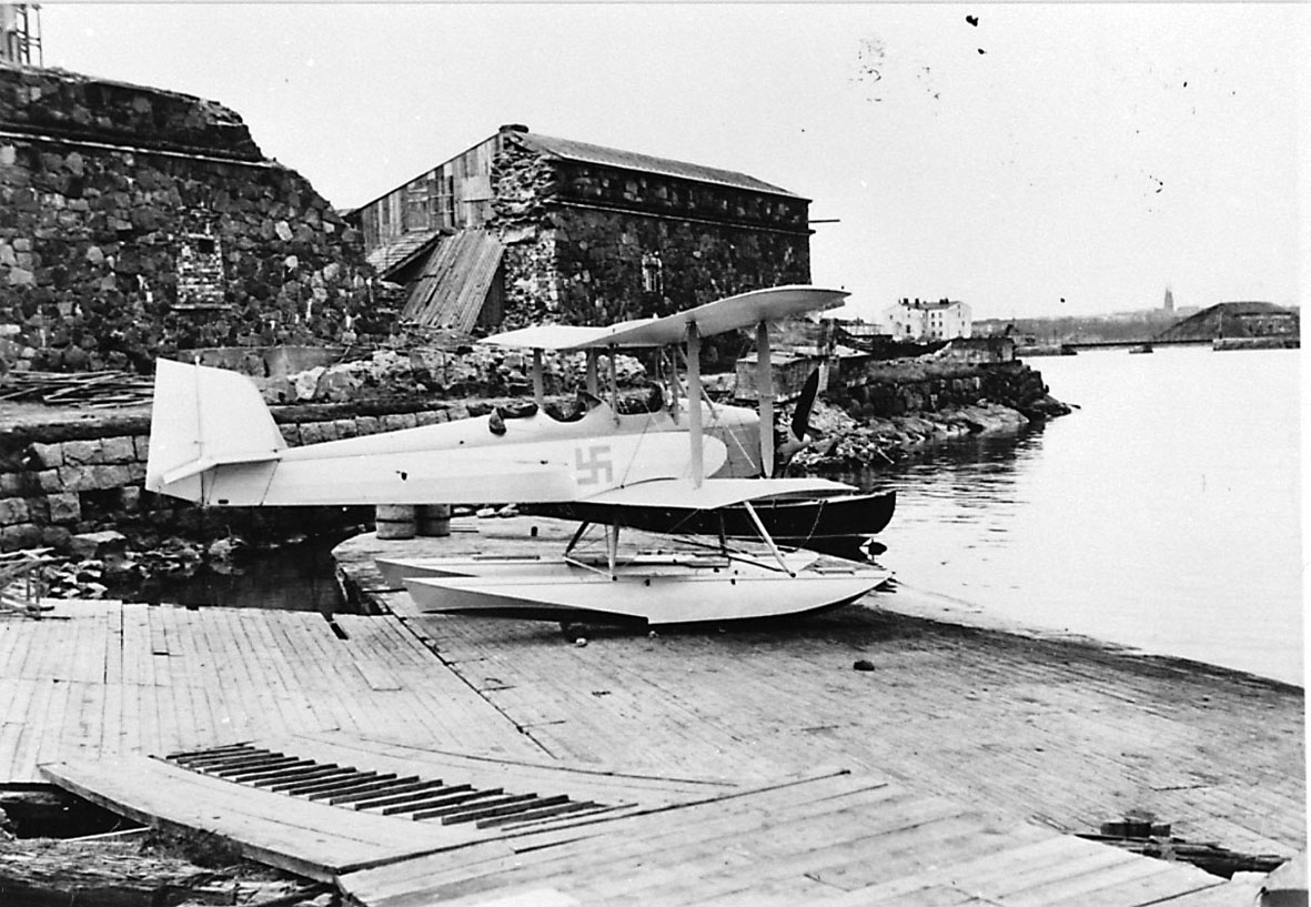 Fly, propellfly, dobbeldekker, Sääski II, med hakekors symbol på siden. Sjøfly som står på land nede ved havneområdet.