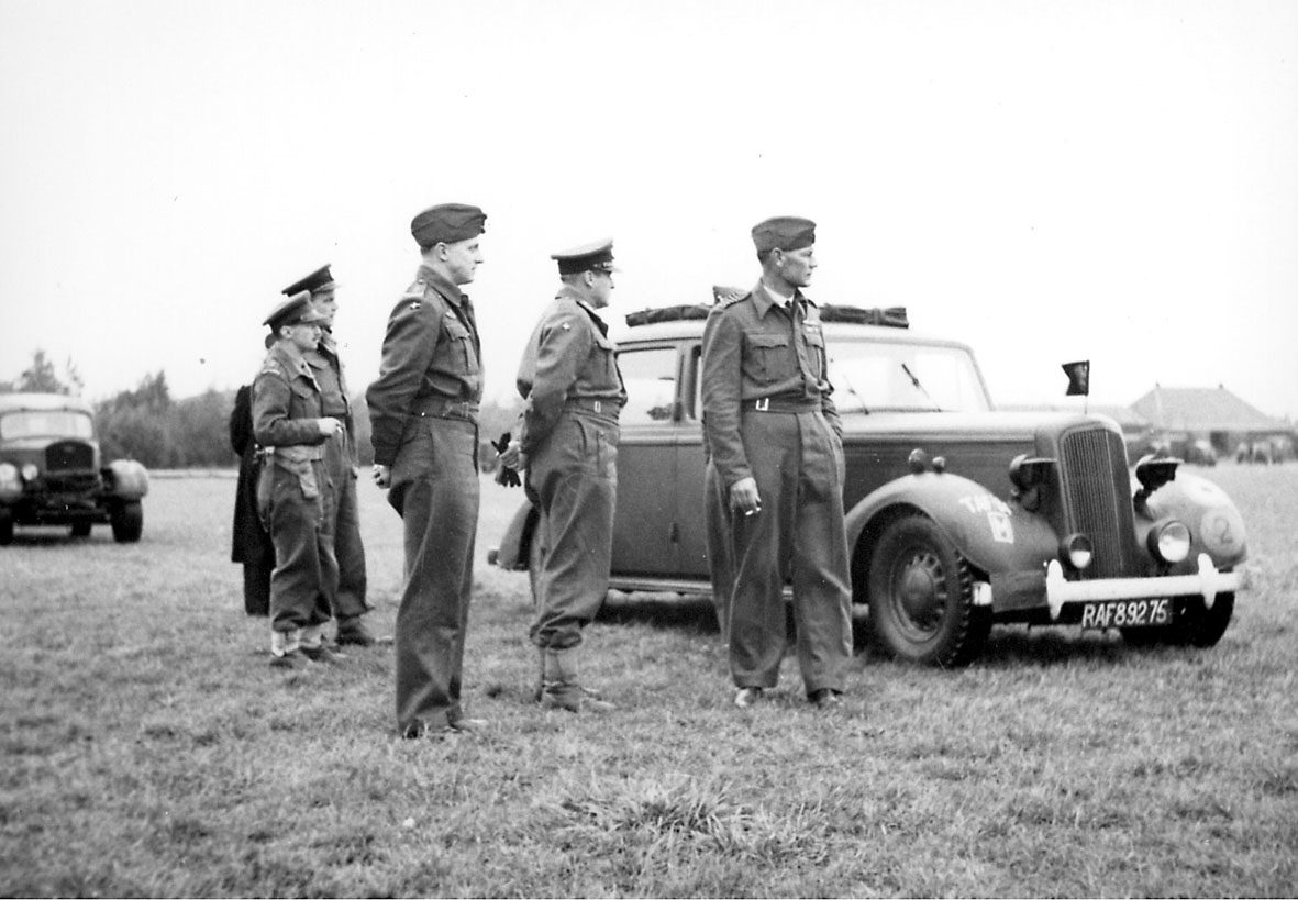 6 personer i militæruniform. Kronprins Olav sammen med 5 andre, på åpen plass. 2 biler, foran med nummerskilt RAF 89275