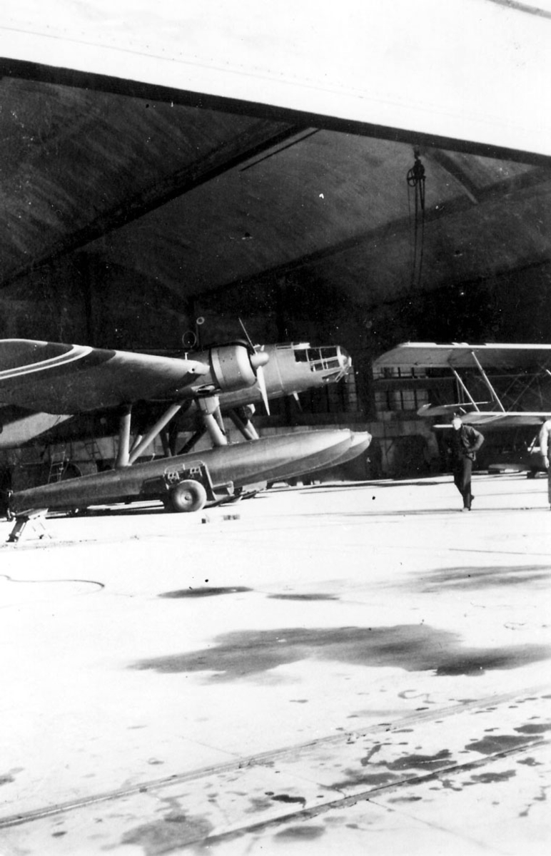 1 fly på bakken, ant. inne i hangar, Heinkel He 115 (N) A 2. Litt av annet fly t.h., dobbeldekker. 2 personer