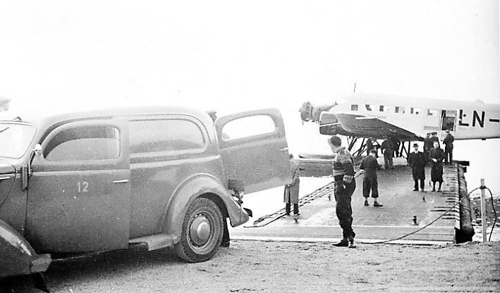 Sjøflyhavn, 1 fly, Junkers ligger ved flytebrygge. Flere personer og kjøretøy, Ford V8 1937 varebil.