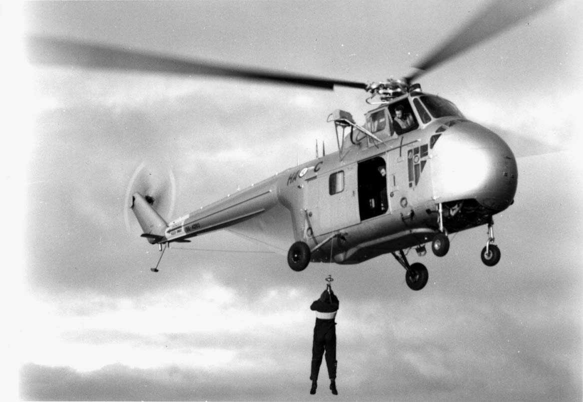 Lufthavn - flyplass. Ett helikopter Sikorsky H-19  Chickasaw   HA-C  like over bakken. En person henger i en line - vaier fra helikopteret.