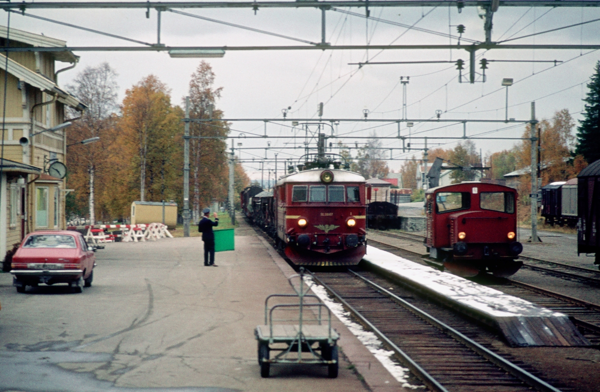 Raufoss stasjon. Togekspeditøren viser signal "Passer" til godstog med El 11 2087. I spor 2 står en skinnetraktor type 214 som har kommet med kipptog fra Gjøvik.