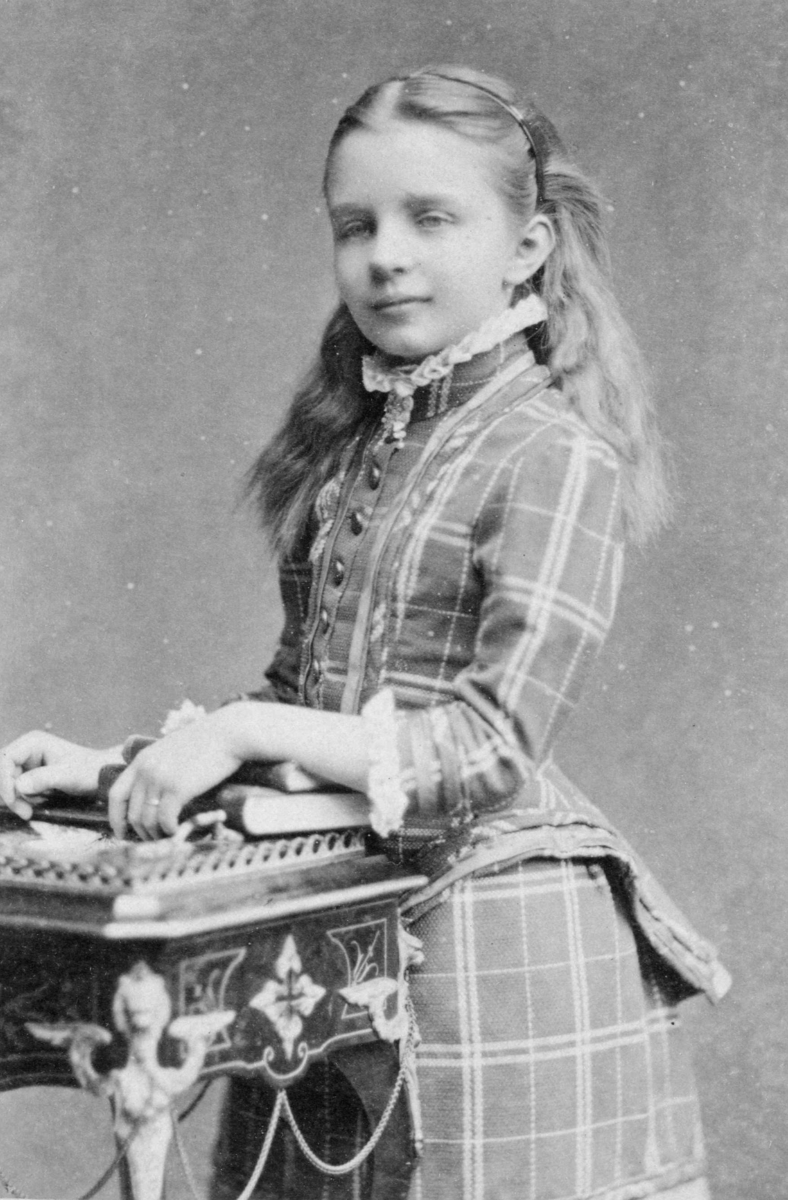 Thea Schweigaard i rutet kjole med kø og høy hals, ca. 1883