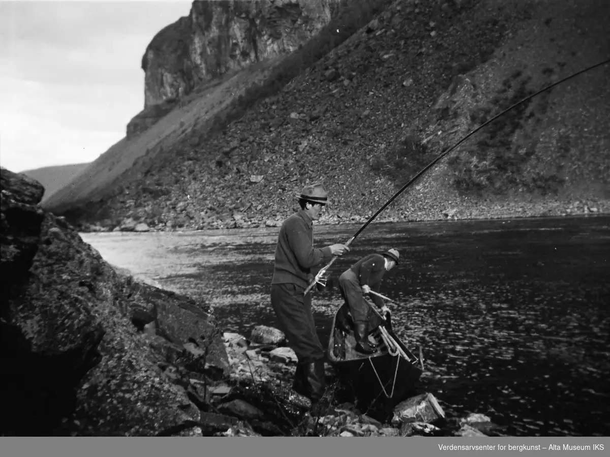Paul Tangen fisker i elva, og en mann står i elvebåten.
Bildet er tatt i fiskesesongen på sommeren i 1949.