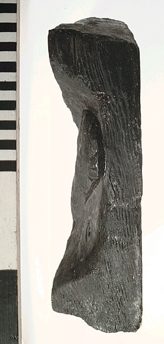Del av skulptur. Rektangulär form, med snedskurna kortändar. Den ena långsidan är avfasad.  Avsmalnande i ena änden. Ornamenterad och profilerad framsida, slät baksida. I den smalare änden syns ett runt hål, med spår efter spikhuvud.
Skulpturdelen är relativt sliten samt bruten i ett hörn.