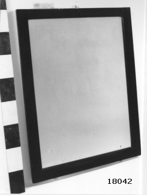 Rektangulär spegel med slät träram av betsad och fernissad mahogny, ramens B = 50 mm. Baksida klädd med kartong. Plattor för fastskruvning mot skott.