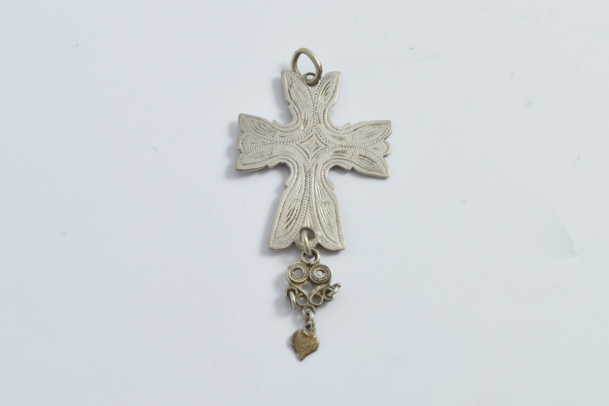 Korsformet anheng i sølv. Anhenget er gravert med bladlignende ornamenter. På undersiden av korset er det festet en filigransdobbe med hjerteformet løv.