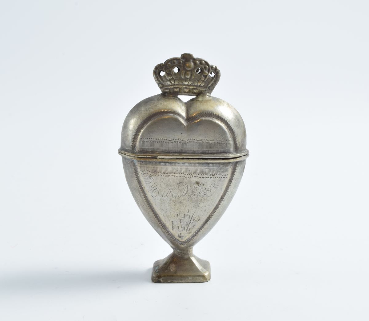 Hjerteformet på firkantet fot, krone på lokket, riflet horisontal bord på hus og lokk, gravert bladornament i hjertespissen.
