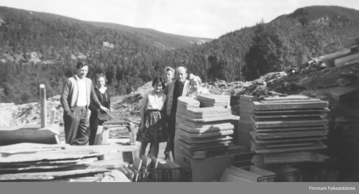 I skiferbruddet i Alta i 1962. Fritz Ebeltoft holder armen rundt datteren Torill. De øvrige er ukjente. Vi ser store stabler med skiferheller i forkant av bildet