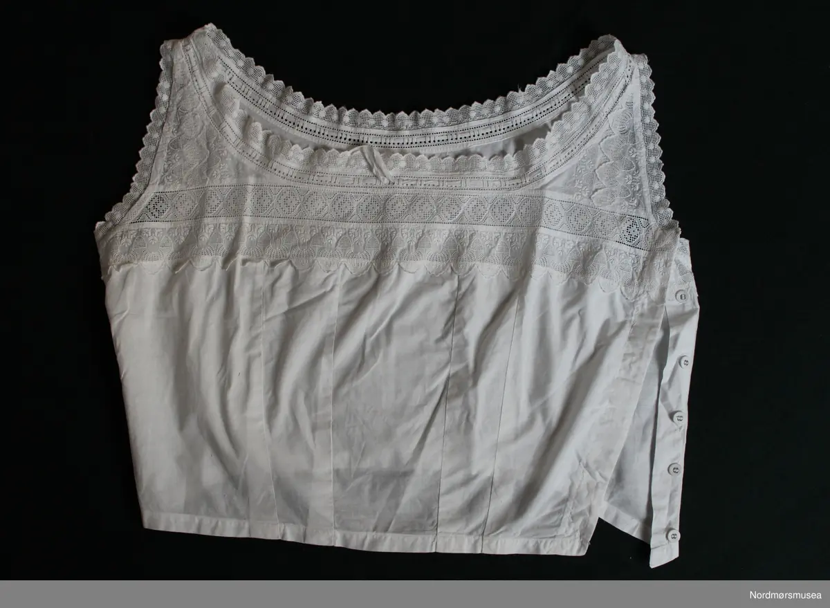 Kort liv. Undertøy til dame. Figursydd. Knapper i venstre side, 6 stk. Innfelte blondebånd, fra brystet og opp. Knytebånd ved avslutning mot hals.