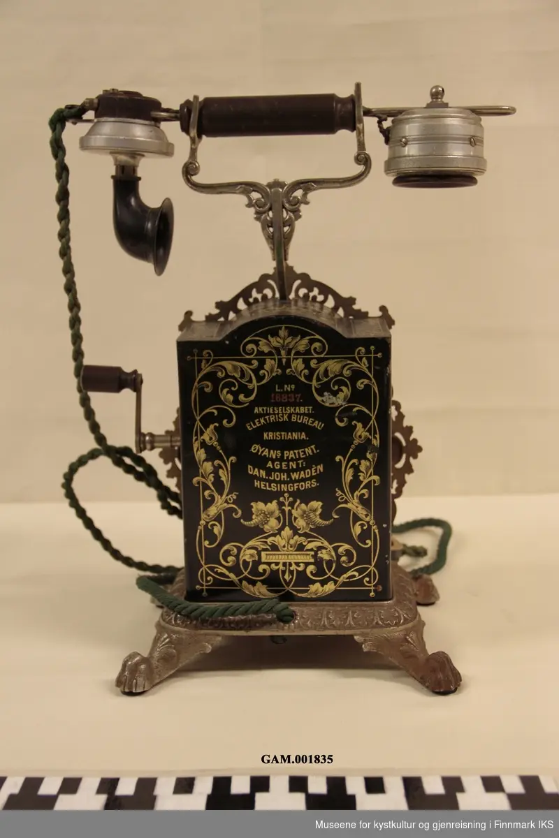 Telefonapparat m/ sveivehåndtak på høyre side. Buet front av støpejern og ben formet som løvepoter. Dekorert med akantusranker.