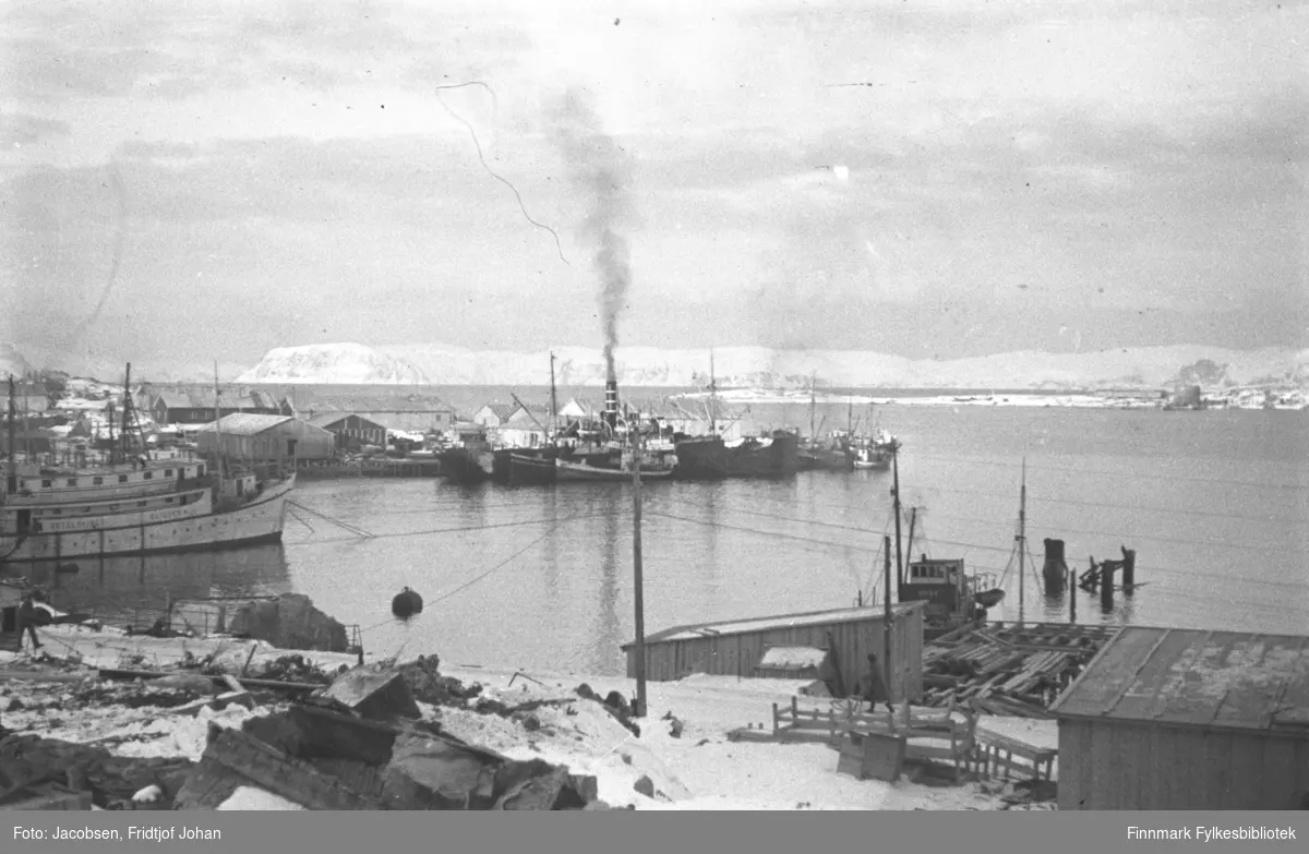 Havna i Hammerfest. Hospitalskipet Elieser 4 ligger lengst til venstre i havna. Flere andre båter ligger ved kaia og endel havnebygnine ses på Hammerfest-neset. Et par brakker ses nede til høyre på bildet og en kai er under bygging. Båten som ligger senket i sjøen til venstre på bildet er FFR-båten Brynilen som ble senket ved kai under krigen. En stabel med ruiner etter et bygg ligger i terrenget nede til venstre på bildet. Det er endel snø på området og over den blikkstille havna ses Fuglenesodden. Fjellene i bakgrunnen er Sørøya.