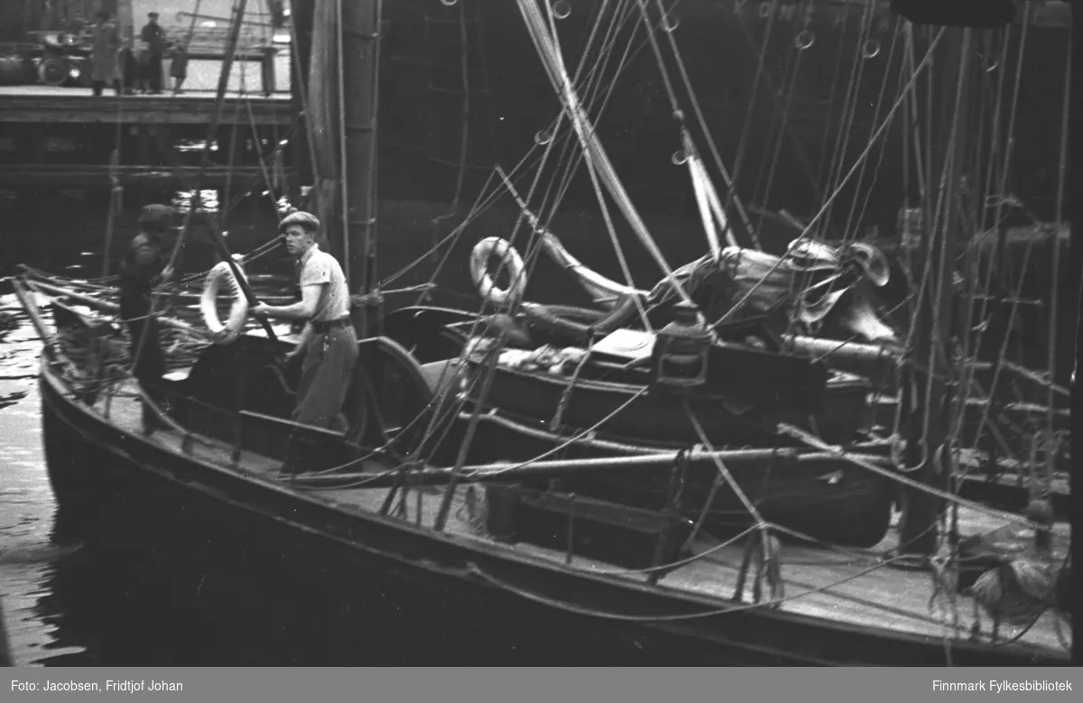 En seilbåt på vei til kai i Hammerfest og den passerer hurtiguteskipet Kong Harald. En mann står på fordekket med en båtshake i hendene. Båten har mye tauverk som går på kryss og tvers fra masten og til rekka. Oppe til venstre på bildet ses en kai med noen mennesker oppå.