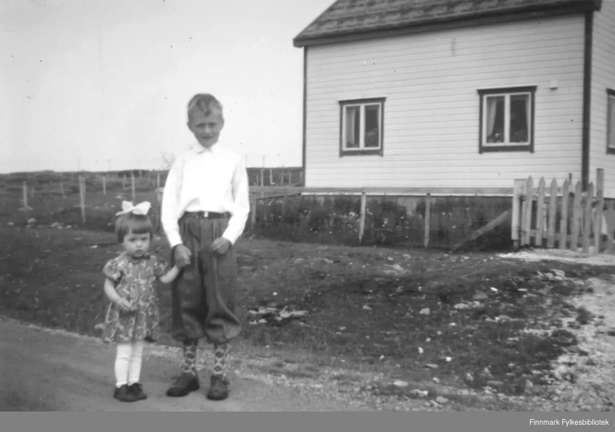 Tormod og Svanhild Rushfeldt poserer foran et hus. Storebroren holder lillesøster i handa. Sommerlig vær skal man dømme etter påkledningen. Hun i mønstret sommerkjole og sløyfe i håret. Han i hvitskjorte og knebukse.