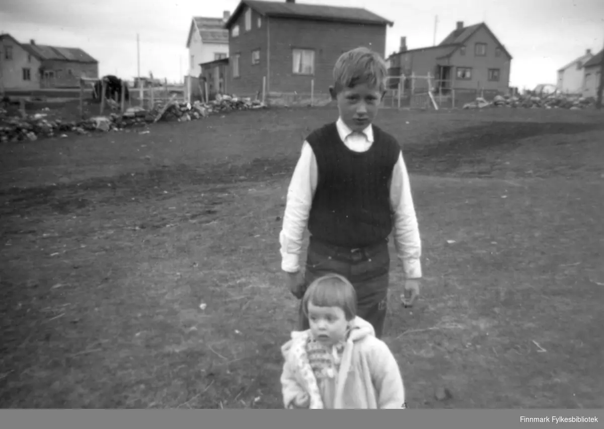 Søskenbarnene Øystein og Svanhild Rushfeld fotografert på en slette foran 5 hus. Hun kledt i en lys, ganske stor jakke, han i olabukse, hvitskorte med slip-over. Flere av husene er i dag revet.