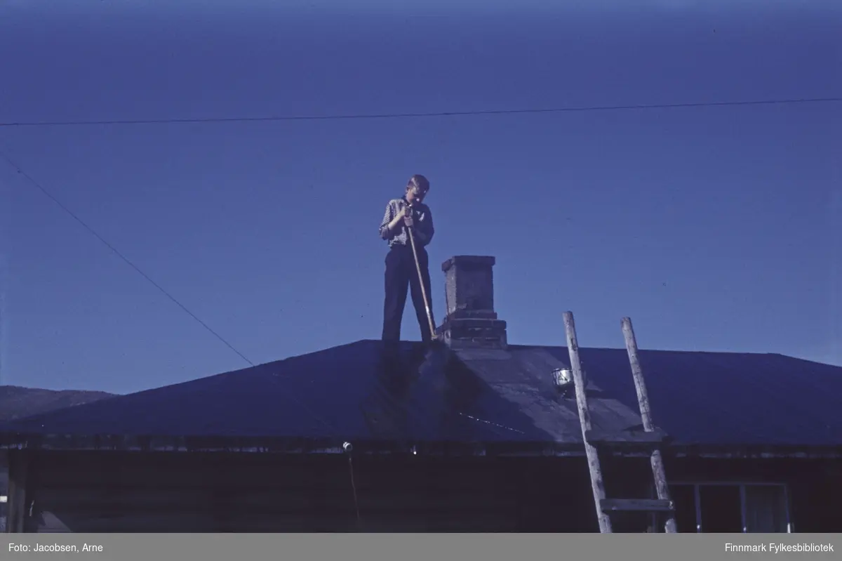 Arne Jacobsen smører tjære på et valmet tak med pappdekke. Han står på takmønet ved siden av teglsteinspipa og har en tjærekost med langt skaft i hendene. På seg har han en mørk bukse og en lys skjorte med ermene oppbrettet. En hjemmelaget trestige står oppreist mot taket og et spann henger fra ene stolpen. Bygget har liggende panel og et tredelt vindu  med gardiner ses på langveggen. Mot den blå himmelen går to kabler, en på tvers og en ned mot bygget.