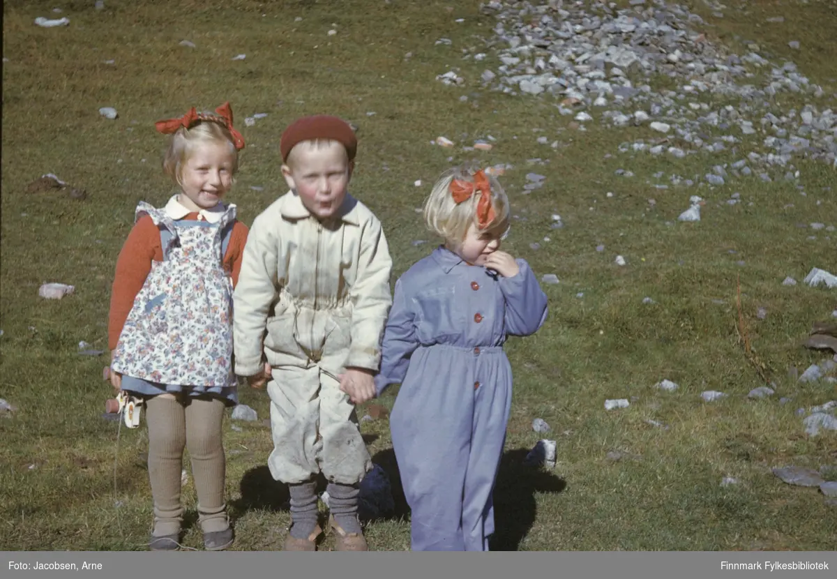 Arne Jacobsen fotografert sammen med to ukjente, små jenter som han holder i hendene. Han har en lys buksedress, lue og store sko på seg. Jentene har sløyfe i håret og hun til høyre på bildet har  en posete buksedress på. Jenta til venstre har strømpebukse, langermet genser og forkle. Sola skinner og en del stein ligger i terrenget.
