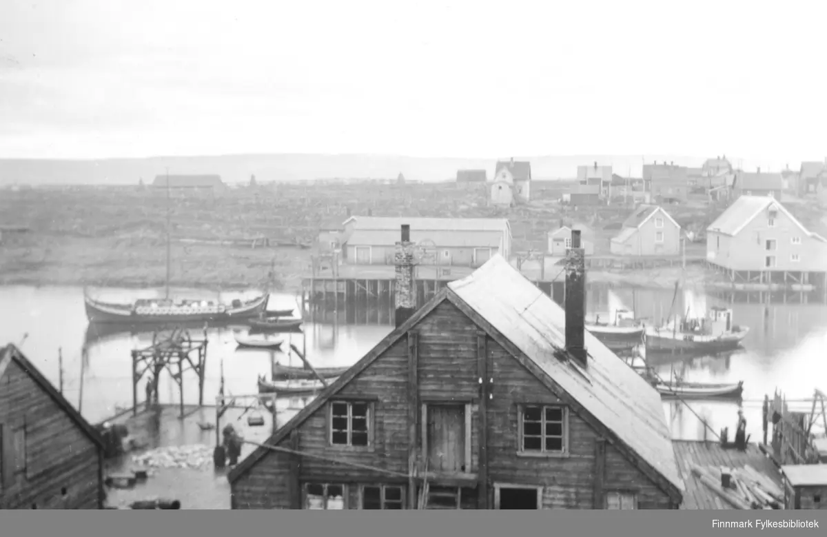 Fotografi fra Vardø. På bildet ser man utover havna i Vardø. Det ligger en del båter på det stille vannet. Til venstre i bildet ligger det en båt som ser ut som et vikingskip. Det er mange skjold på siden av båten. På kaien er det noen mennesker