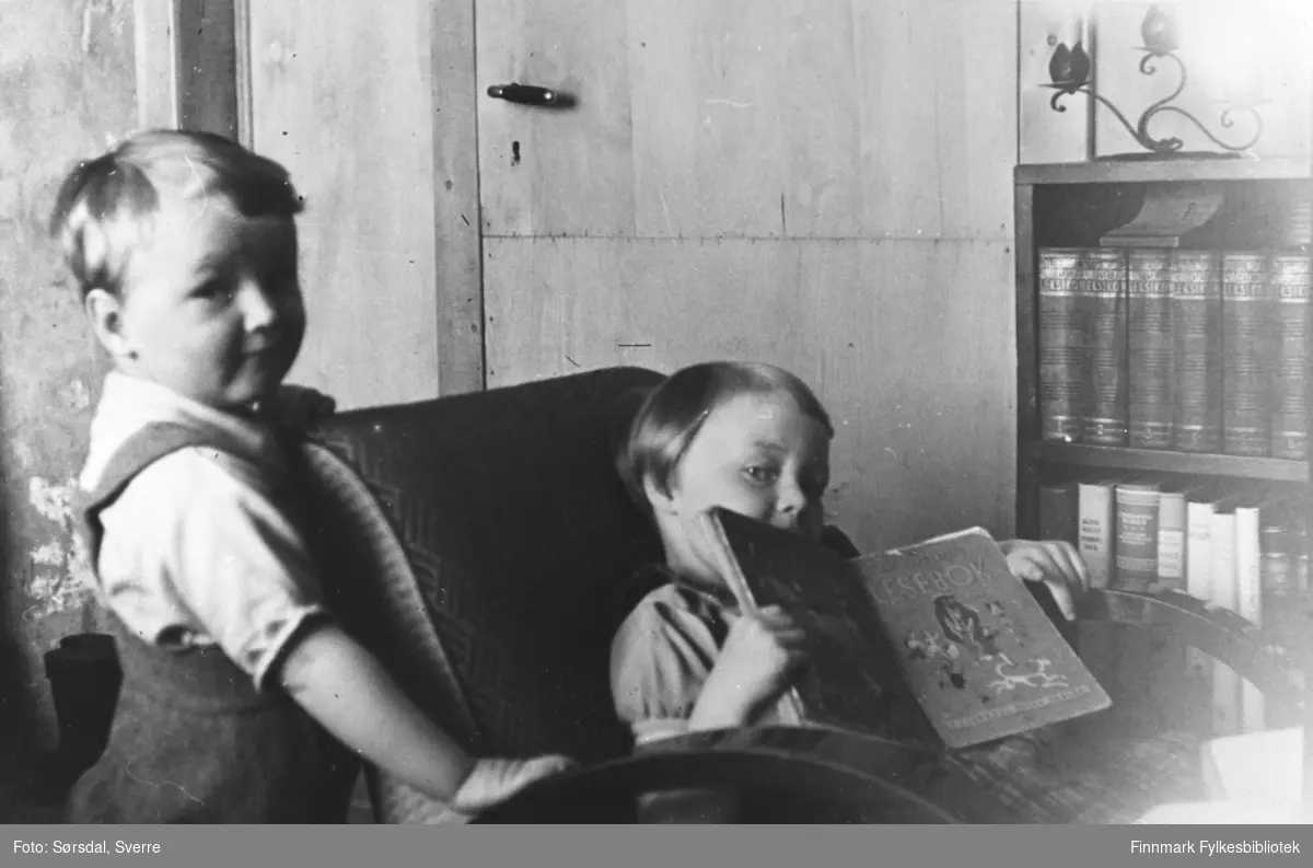 Eirik og Randi Sørsdal i Syltefjord. Jenten sitter i en stol og ser i en bok mens gutten står ved siden av. Ved siden av stolen står en bokhylle og vi se døra i bakgrunnen.
