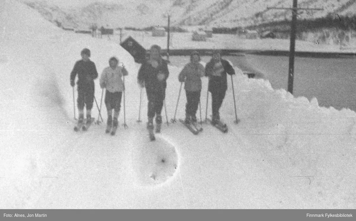 På ski gjennom Vassdalsura i Øksfjord, vinteren 1955. Fra venstre: Ingolf Bye, Torfinn Kiil, Evy-Ann Larsen, Hildegunn Hansen, Karin Mikkelsen. Det store huset i bakgrunnen var det såkalte 'nothuset', som lå der dagens fergeleie er