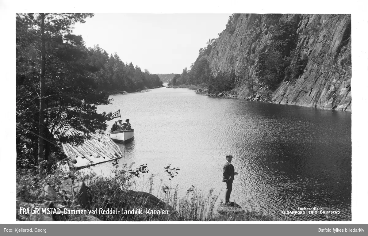 Dammen ved Reddal-Landvik-kanalen, Grimstad i Aust-Agder. Båt med vimpel med teksten STRI...?o kvinner, en mann og to barn ombord. 
Mann i forgrunnen. Tømmer i vannet.