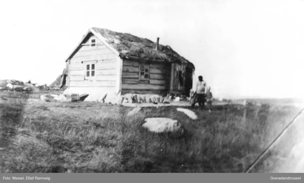 Mann med to barn foran tømmerhus med torvtak