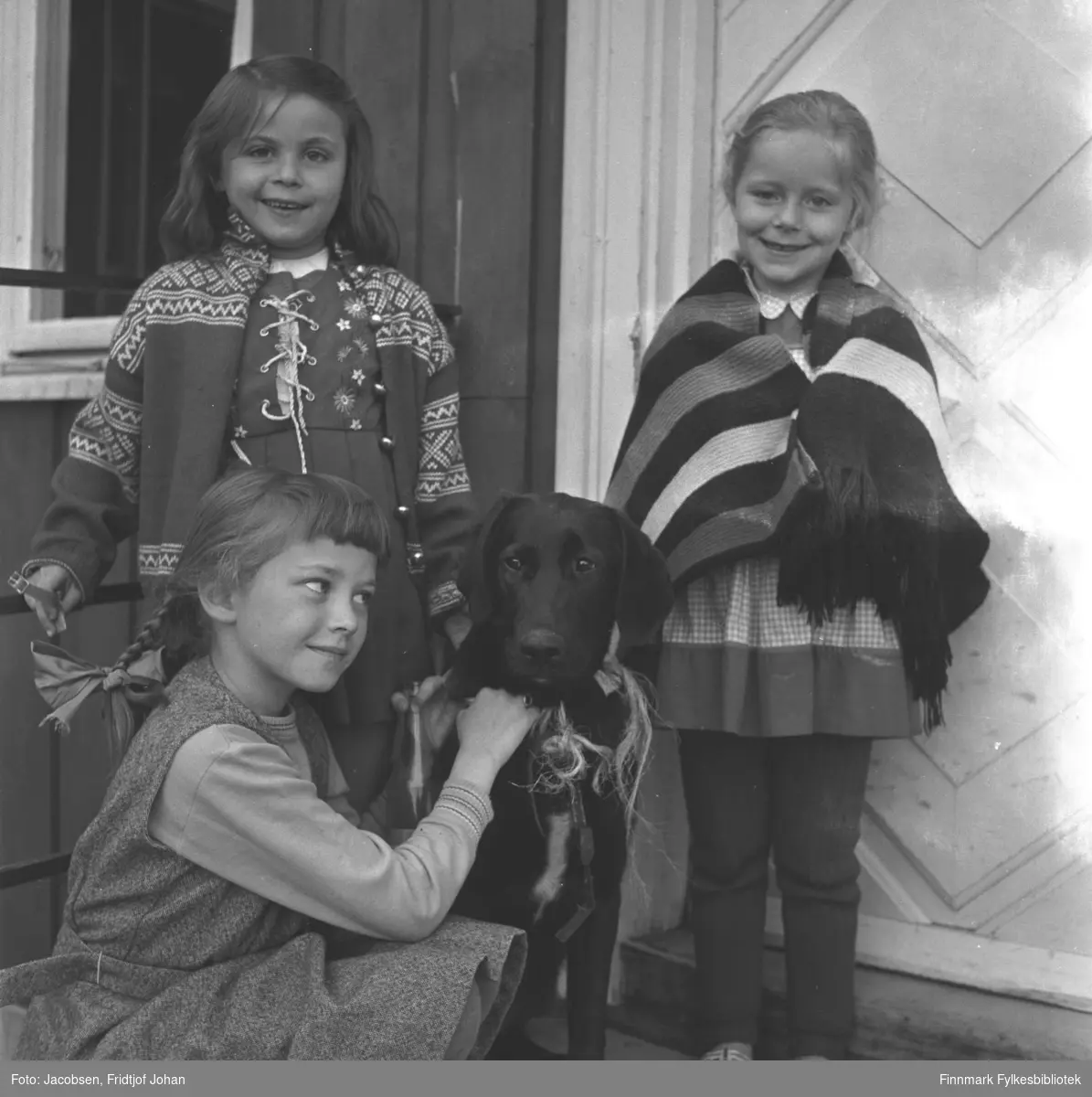 Tre små jenter og en hund fotografert på trappa til et hus. Jentene og nøyaktig sted er ukjent.