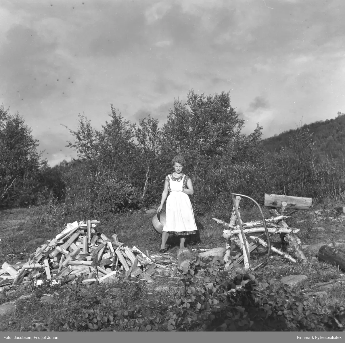 En kvinne står rett bak en hjemmelaget sagstol og en haug med ved.