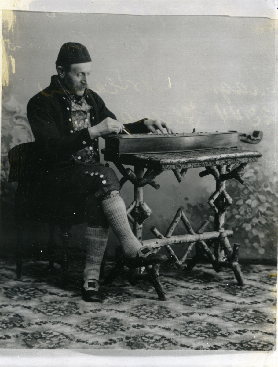Langeleikspelaren Ola Brenno, i folkedrakt med langeleik, fotografert med lerretbakgrunn innandørs