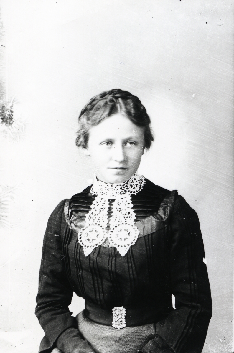Ung kvinne avbildet i svart bluse og grått skjørt. Hvit, heklet krage og pyntet beltespenne.