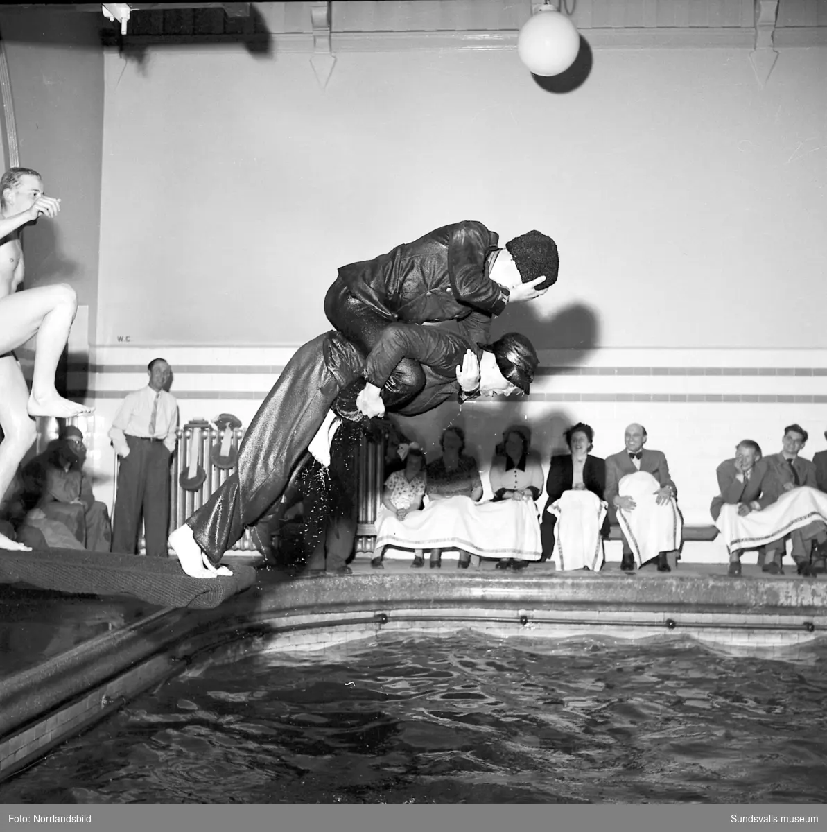 Simtävlingar i gamla badhuset vid Kyrkogatan. Även simhopp av det spexigare slaget. Notera att publiken försetts med handdukar över knäna för att skydda kläderna mot stänk.