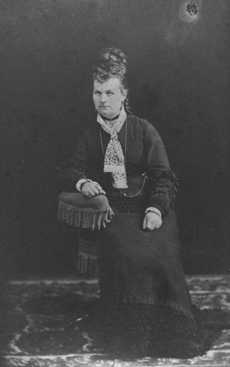 Visittkortportrett av en russisk kvinne med 'høy' frisyre. Kvinnen sitter på en lenestol med armen på ryggstøet. Stolen, bakgrunnen og gulvet er samme som på bilde fb.96009-020
