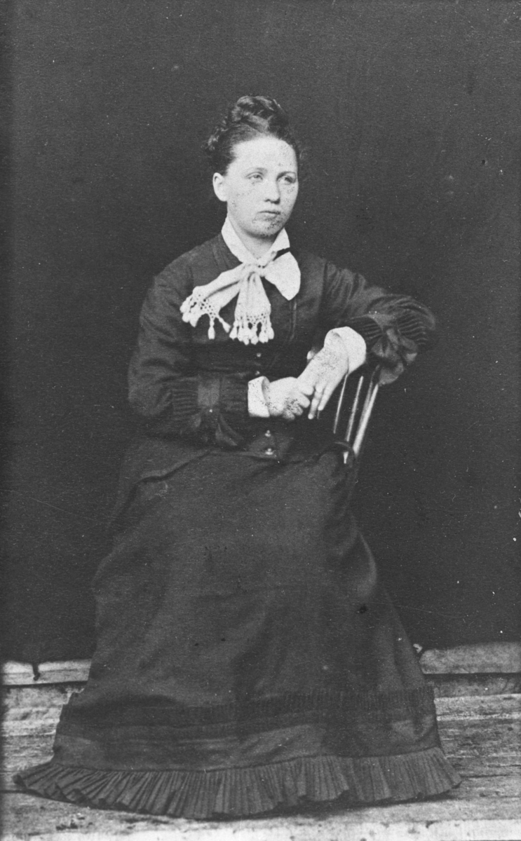 Portrett av en kvinne i mørk lang kjole, skjorte og en stor hvit sløyfe rund halsen. Hun sitter på en stol og lener med venstre armen på ryggstøen. Dette visitkortet har døde kanter (P.O.Næss?) og det er tatt tidlig på 1860 årene, kanskje tidligere