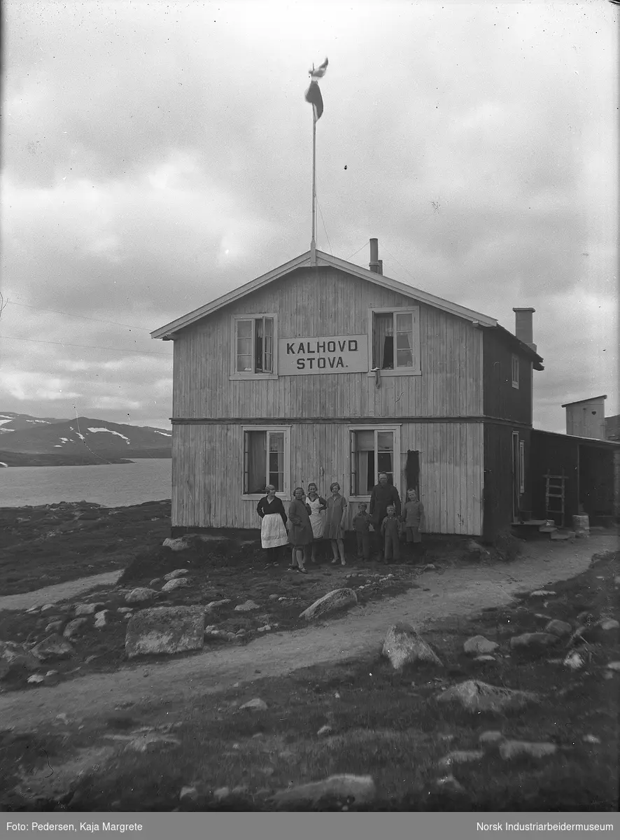 Mennesker oppstilt foran Kalhovdstova. Flaggstang på byggets møne der det norske flagget er heist. Toalettbygg på høyde bak bygget.