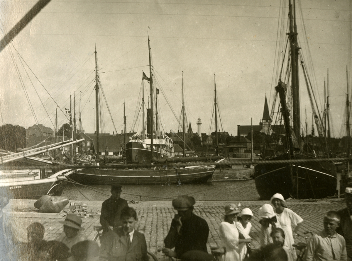Diverse människor samt segelfartyg. På fartyget längst till vänster finns texten VESTER skriven på skrovet. I bakgrunden finns även en passagerarbåt.