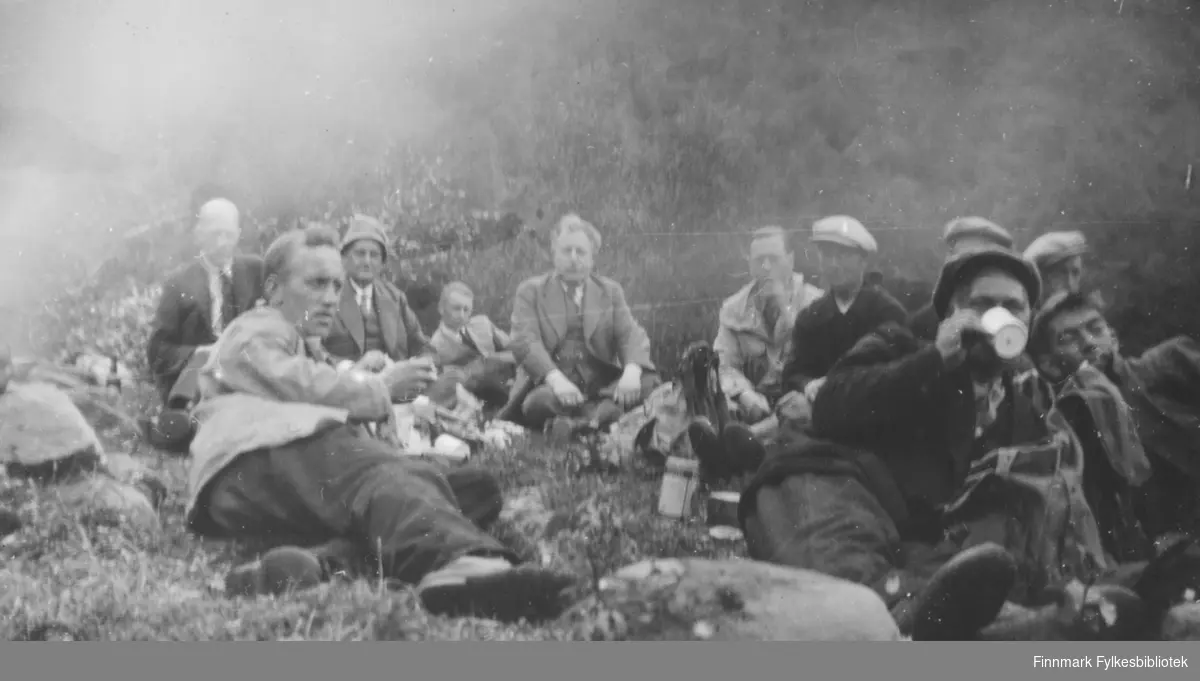Elvebåtstakere sitter samlet i bakken, trolig for en matpause. Flere menn i dress og slips er med. Antagelig 1930 tallet.