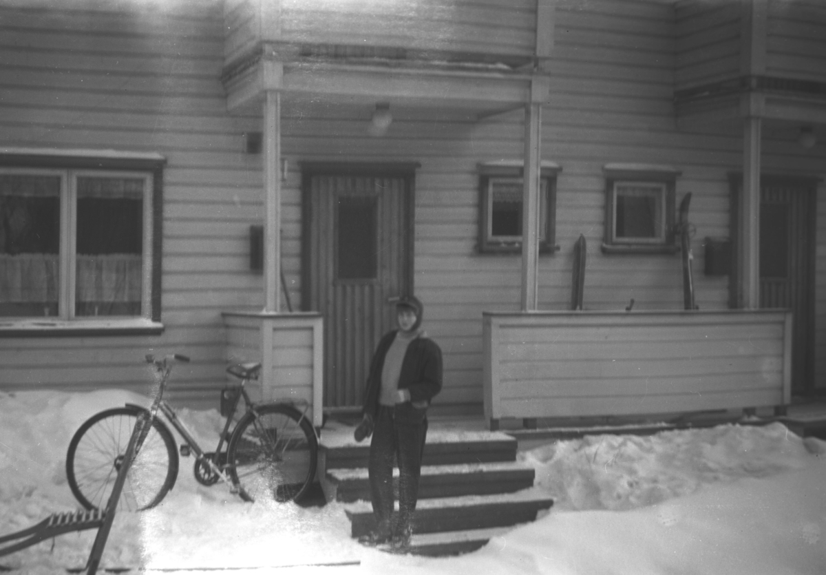 En ukjent, ung person står ved inngangspartiet til et hus. Både en spark og en sykkel står i snøen. Stedet er ukjent.