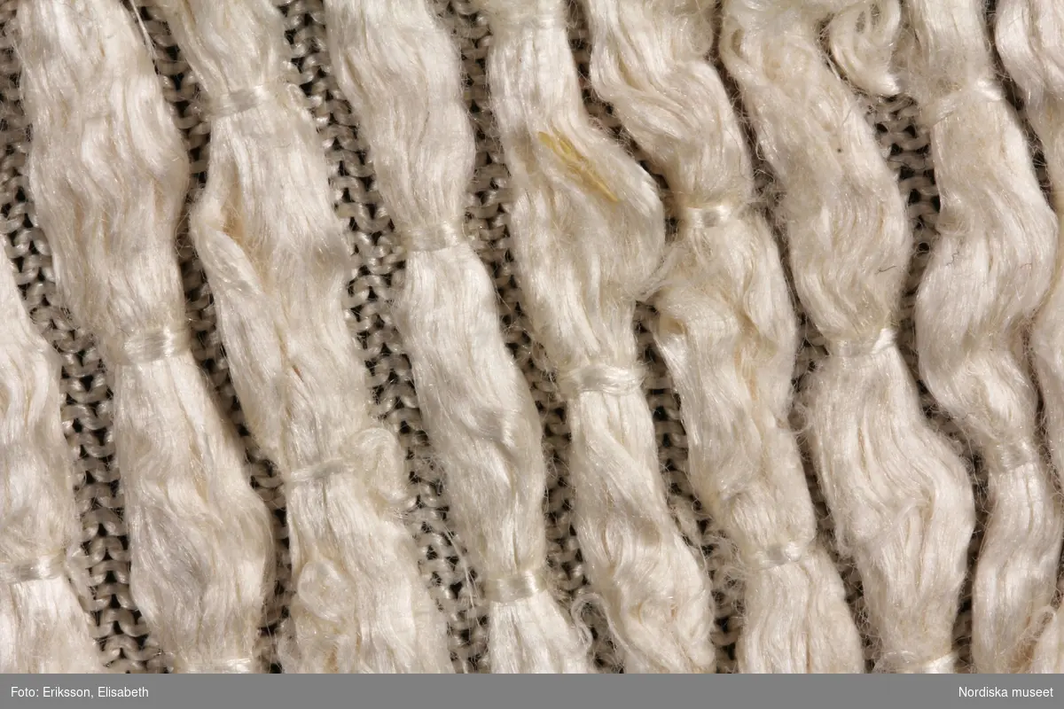Katalogkort:
Kofta
för dam, hvit silke

L. i ryggen omkr 0,60m.
Af hvit trikåväfvnad i silke med på afvigsidan löst inväfda tjocka silkestrådar. Dessa trådar äro på jämna afstånd fästade i väfven så att rätsidan verkar prickig. Kring halsen ett dubbelvikt grönt och hvitt sidenband. Koftan sammanhålles med tre par hvita sidenband, som knytes. Består af två ryggstycken och två framstycken med kilar i sidorna i skörtet. Armarna halfkorta, med endast en söm.