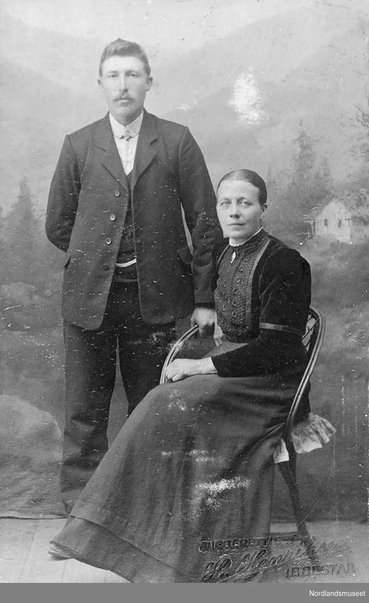 1 kvinne og 1 mann i helfigur. Kvinnen sitter på en stol mens mannen står ved siden av. Mannen er kledd i mørk bukse, mørk jakke, mørk vest, lys skjorte og lyst slips. Kvinnen er kledd i mørk lang kjole. I halsen på kjolen er det festet en nå. Personene på bildet er Olaf Jentoft og Marie Olsen (Olafs tante), Torpelvann.