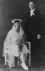 1 brudepar. Bruden sitter i en stol. Hun er kledd i en lys k