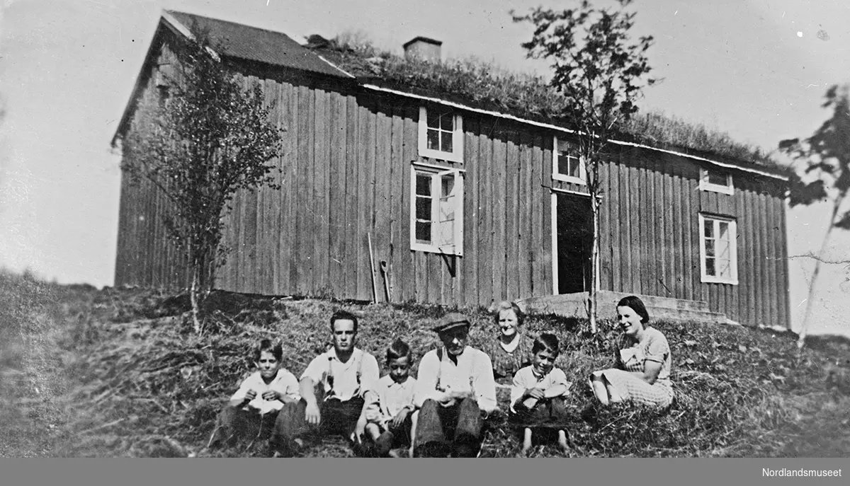 3 menn, 2 kvinner og 2 barn som sitter på marken utenfor et hus.