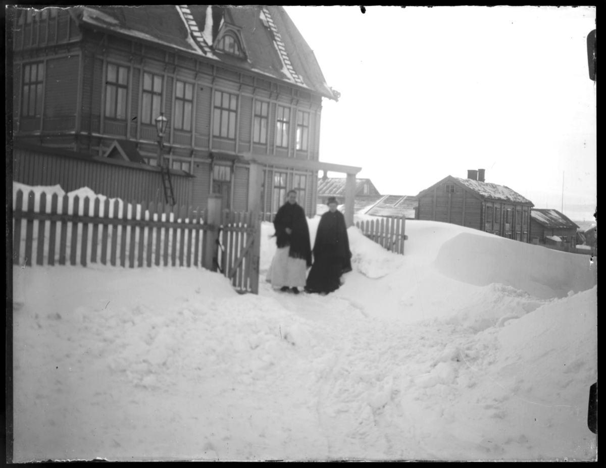 To kvinner står i snøen i det som ser ut til å være porten inn til prestegårdshagen. Den store bygningen i bakgrunnen er skolen i Vardø sett fra en av kortsidene. I bakgrunnen et hus som ser ut til å være et vanlig bolighus