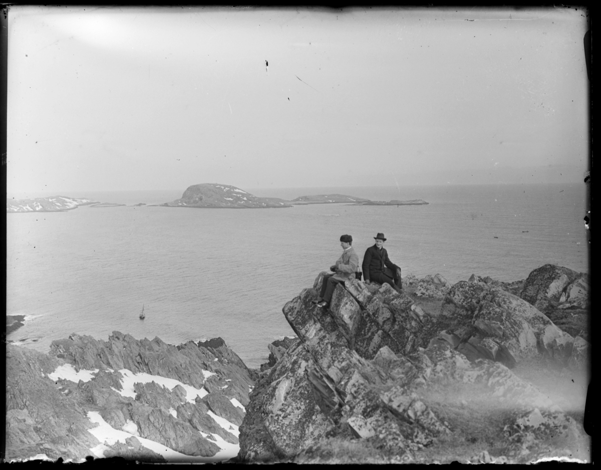 To menn sitter i et klippeframspring på Rødmoen med utsikt over havet og Reinøya og Hornøya. En seilbåt ligger fortøyd nedenfor klippeutspringet