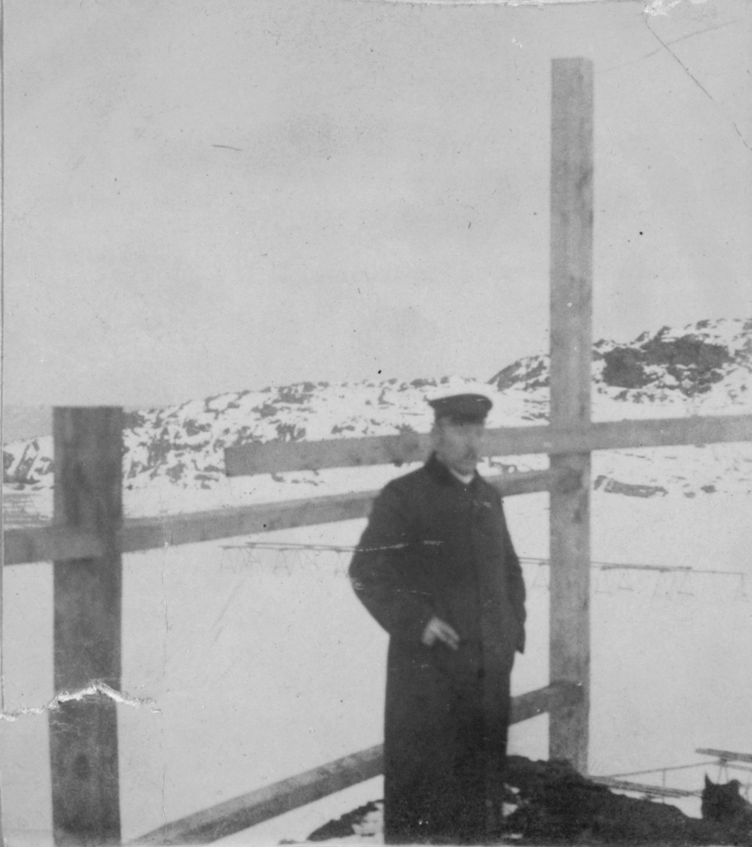 Karl Ohlsen i hatt og frakk. Han står ved noen stolper eller gjerde. Det er snø på marka.