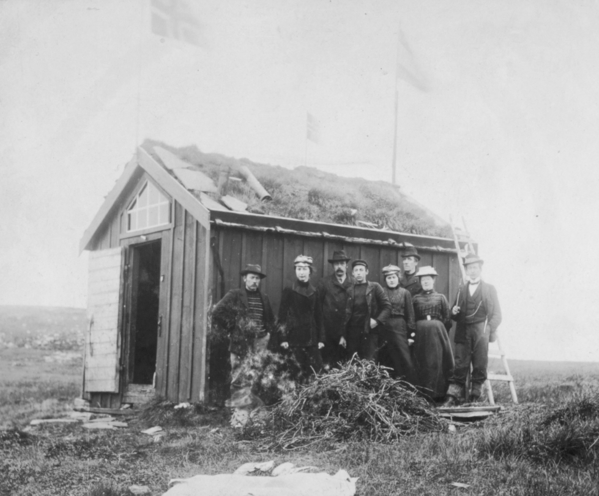 August 1904. Damer og menn står oppstilt utenfor en hytte. Det er torv på taket. Hyttedøra står åpen. Flagg på taket.